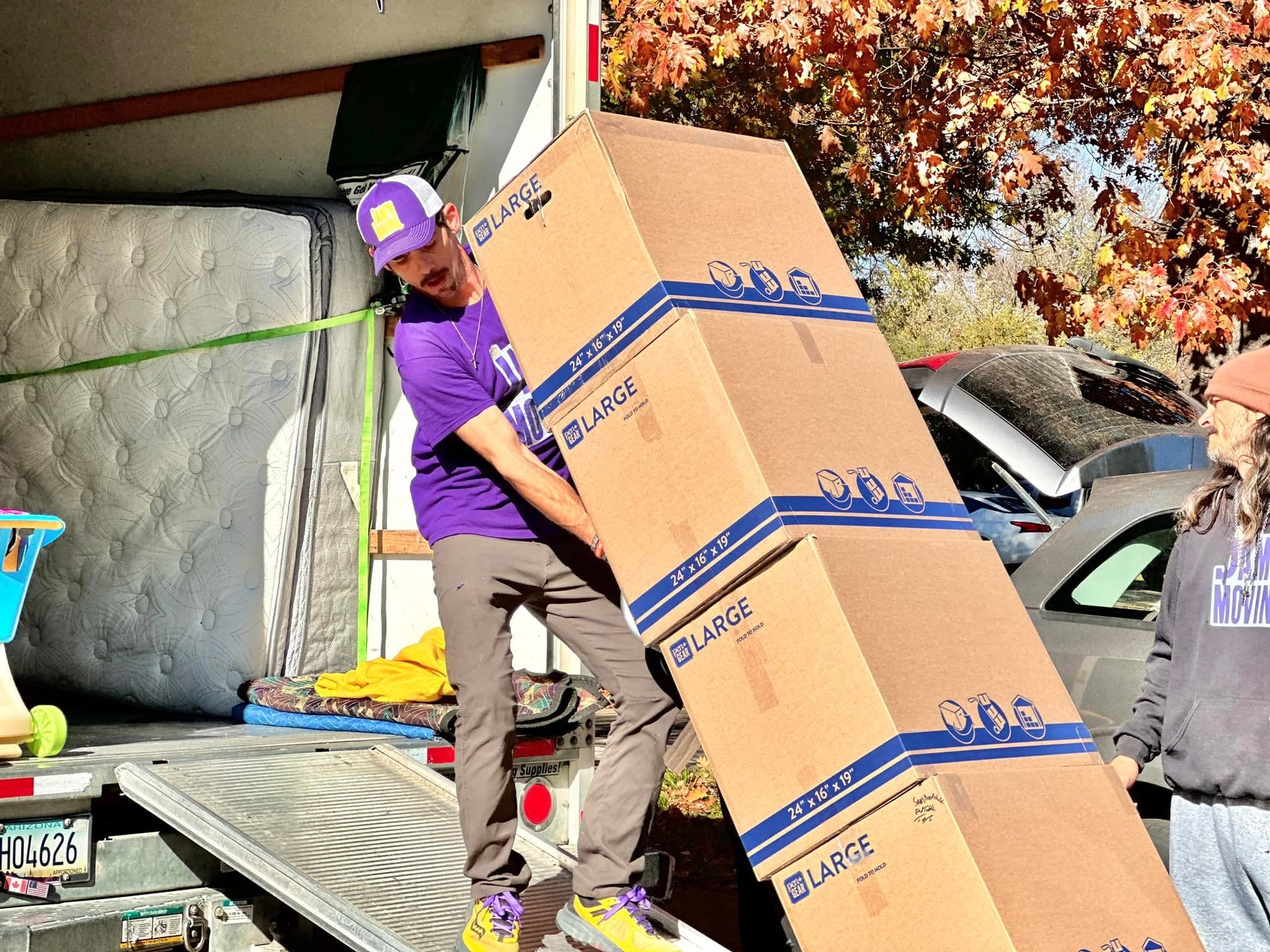 A Jam'N Moving crew member carrying a stack of large moving boxes down the truck ramp